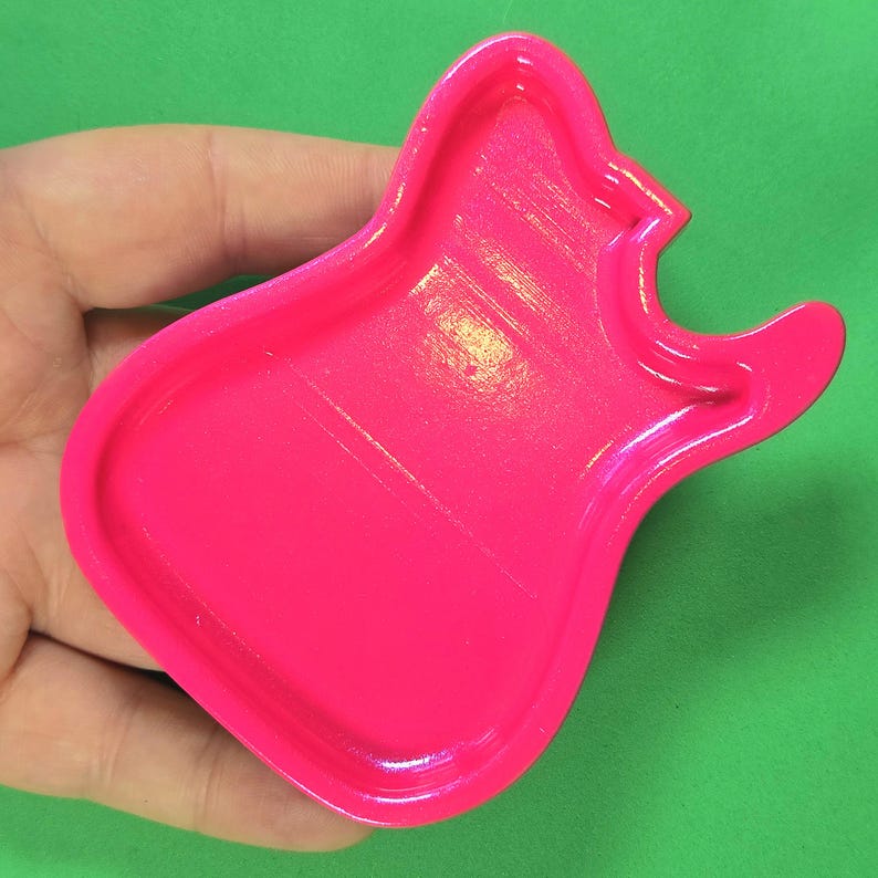 May include: A bright neon pink guitar-shaped cookie cutter with a raised rim and a recessed center. The object features a distinct body shape with a cutout neck and headstock area, designed for cutting dough into the silhouette of an electric guitar.