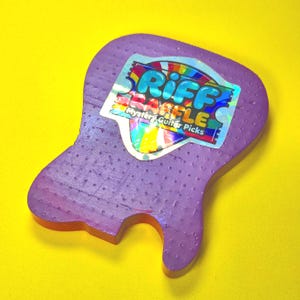 May include: A purple, guitar-shaped object with a textured surface featuring a holographic sticker in the center. The sticker displays the text "RIFF RAFFLE" in blue letters above the words "Mystery Guitar Picks" in white, set against a rainbow background.