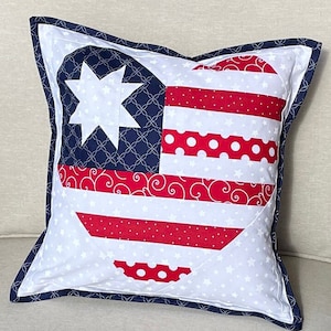 May include: A white pillow with a blue trim features a red, white, and blue heart-shaped design with a star in the upper left corner. The heart is made up of various patriotic patterns, including stripes, polka dots, and swirls.