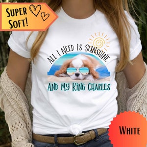 May include: White t-shirt with a graphic of a Cavalier King Charles Spaniel wearing sunglasses and laying on a beach with the text "All I need is sunshine and my King Charles".