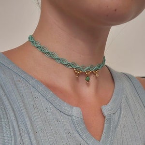 Op de afbeelding: Een lichtblauwe chokerketting met een gevlochten ontwerp en een hanger van goud en groen.