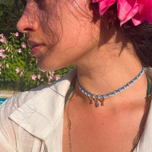 May include: A choker necklace featuring a light blue and white woven band with a scalloped edge, accented by three small gold-toned charms with a central blue stone, worn with a white collared shirt and a bright pink flower tucked into dark hair.