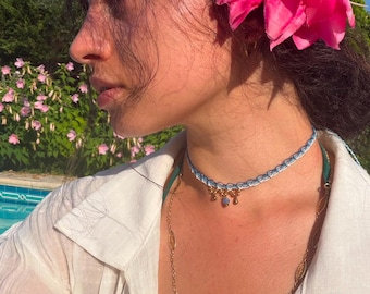May include: A choker necklace featuring a light blue and white woven band with a scalloped edge, accented by three small gold-toned charms with a central blue stone, worn with a white collared shirt and a bright pink flower tucked into dark hair.