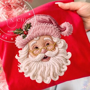 May include: A red Christmas-themed item featuring an embroidered Santa Claus face. Santa wears a pink hat and gold-rimmed glasses, with a white beard and mustache. The item has the text "VILLA N CHICA" and "ORIGINAL DESIGN" in a circular design.