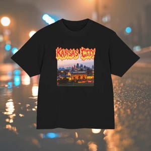 May include: A black short-sleeve t-shirt featuring a rectangular graphic print on the chest. The graphic displays a sunset cityscape of Kansas City with the text Kansas City written above in a stylized, orange and yellow flame-like font.