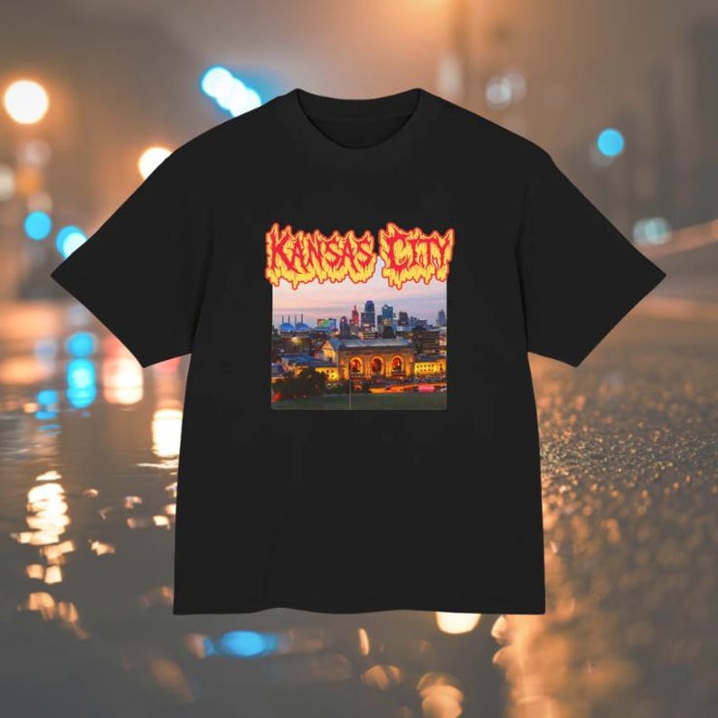 May include: A black short-sleeve t-shirt featuring a rectangular graphic print on the chest. The graphic displays a sunset cityscape of Kansas City with the text Kansas City written above in a stylized, orange and yellow flame-like font.