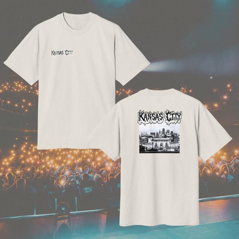 May include: A cream-colored short-sleeve t-shirt featuring the text Kansas City in a stylized black font on the upper left chest. The back of the shirt displays a black and white graphic of the Kansas City skyline with the text Kansas City above.