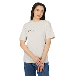 May include: A light gray short-sleeve t-shirt featuring the text Kansas City printed in a black, stylized, jagged font on the upper left chest area. The garment has a crew neckline and a standard fit, constructed from a solid-colored fabric.