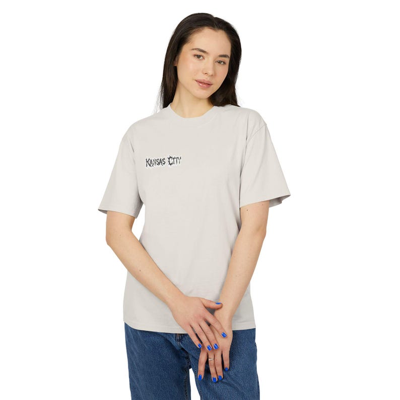 May include: A light gray short-sleeve t-shirt featuring the text Kansas City printed in a black, stylized, jagged font on the upper left chest area. The garment has a crew neckline and a standard fit, constructed from a solid-colored fabric.