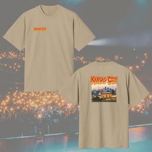 May include: A beige short-sleeve t-shirt featuring a small orange Kansas City logo on the left chest. The back of the shirt displays a larger graphic of the Kansas City skyline with the text Kansas City in an orange flame-style font above it.