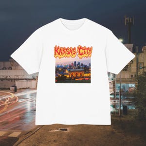May include: A white short-sleeve t-shirt featuring a central graphic print of the Kansas City skyline at dusk. The text KANSAS CITY appears above the cityscape in a stylized, orange and yellow flame-like font against the white fabric background.