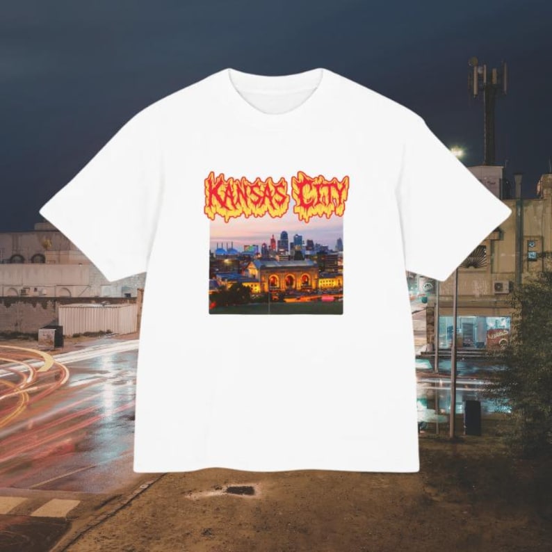 May include: A white short-sleeve t-shirt featuring a central graphic print of the Kansas City skyline at dusk. The text KANSAS CITY appears above the cityscape in a stylized, orange and yellow flame-like font against the white fabric background.