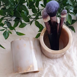 Könnte beinhalten: Zwei zylindrische Behälter, einer mit Make-up-Pinseln. Die Behälter sind weiß mit goldenen Akzenten. Die Pinsel haben dunkle Griffe und violette und beige Borsten. Grüne, belaubte Weinreben sind im Hintergrund.