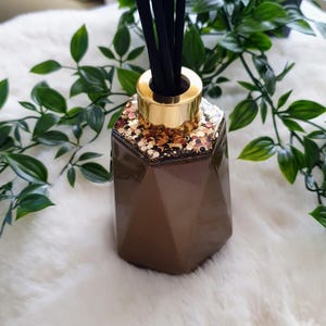 Op de afbeelding: Een bruine, geometrisch gevormde diffuserfles met een goudkleurige bovenkant en zwarte stokjes. De bovenkant van de fles is versierd met goud- en koperkleurige glitter. Groene bladplanten staan op de achtergrond.