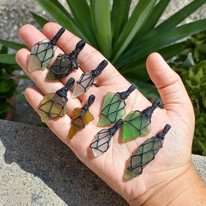 Può includere: Una collezione di otto ciondoli a forma di punta di freccia in vetro di mare, ognuno avvolto in un cordoncino in macramé nero. Il vetro di mare è in varie tonalità di verde, blu e marrone.