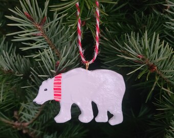 May include: White polar bear Christmas ornament with a red and white striped scarf hanging on a green pine tree branch.