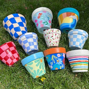 May include: A collection of ten hand-painted terracotta flower pots with various designs. The pots feature checkered, floral, and geometric patterns in shades of blue, red, yellow, white, and orange.