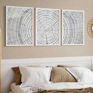 May include: Three white framed prints of black and white abstract wood grain patterns hang above a bed with white and brown bedding. A round wicker mirror with a gold frame hangs on the wall to the right of the prints.