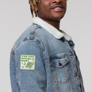 Op de afbeelding: Een lichtblauw denim jack met een sherpa kraag en een groene patch op de linkermouw met de tekst "You are Space Dust" met een vredesteken en een cartoon ruimteschip.