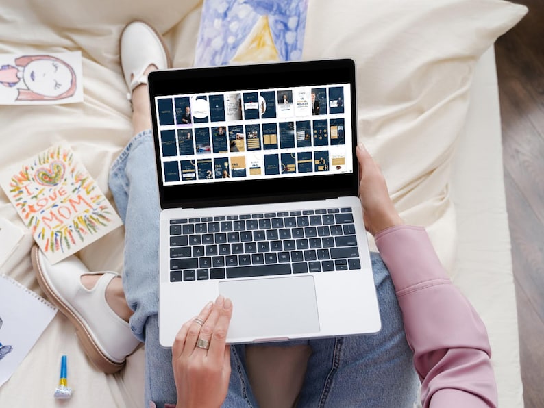 May include: A person is sitting on a bed with a laptop open. The laptop screen shows a grid of small images. The person is using their right hand to scroll through the images. The person is wearing blue jeans and a pink shirt.