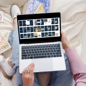 May include: A person is sitting on a bed with a laptop open. The laptop screen shows a grid of small images. The person is using their right hand to scroll through the images. The person is wearing blue jeans and a pink shirt.