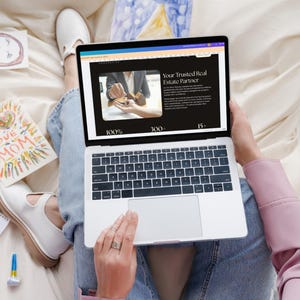 May include: A person is using a silver laptop displaying a website with the text "Your Trusted Real Estate Partner." The person is wearing blue jeans and white shoes. Colorful drawings and a card that says "I Love Mom" are nearby.
