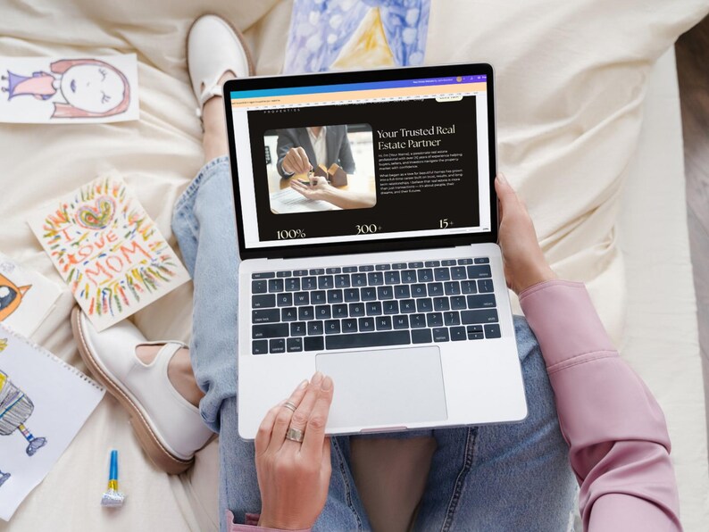 May include: A person is using a silver laptop displaying a website with the text "Your Trusted Real Estate Partner." The person is wearing blue jeans and white shoes. Colorful drawings and a card that says "I Love Mom" are nearby.