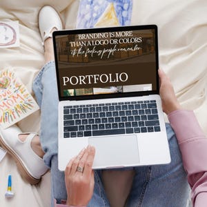 Op de afbeelding: Een laptop toont het woord "PORTFOLIO" en de tekst "BRANDING IS MORE THAN A LOGO OR COLORS is the feeling people remember." Kindertekeningen en witte schoenen zijn zichtbaar.