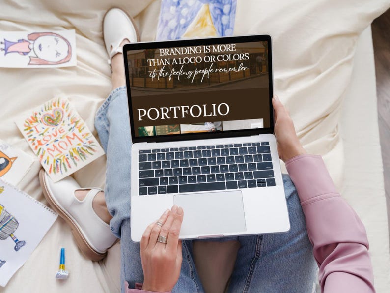 Op de afbeelding: Een laptop toont het woord "PORTFOLIO" en de tekst "BRANDING IS MORE THAN A LOGO OR COLORS is the feeling people remember." Kindertekeningen en witte schoenen zijn zichtbaar.