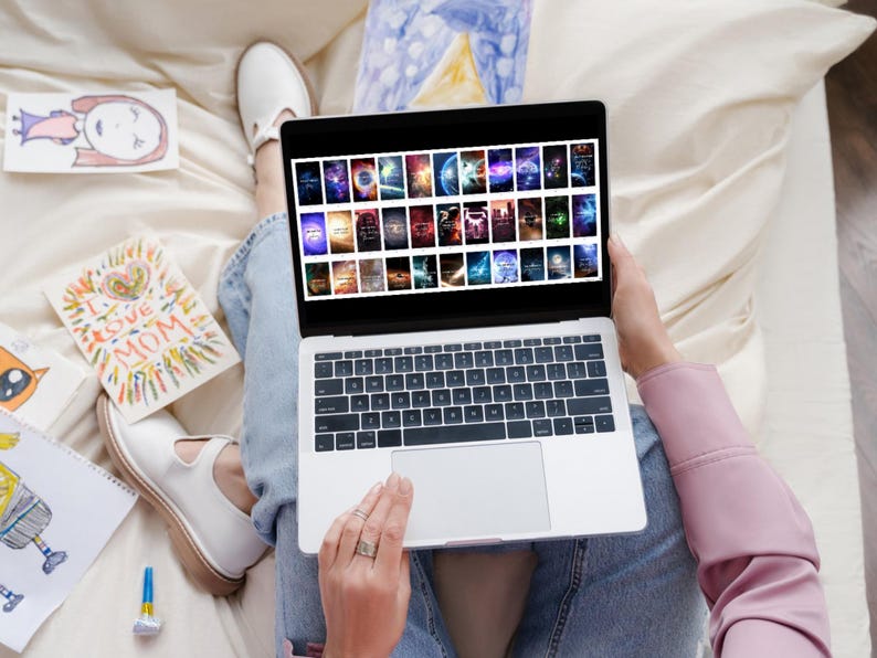 May include: A person is sitting on a bed with a laptop computer open. The laptop screen displays a grid of colorful images. The person is using their finger to scroll through the images.