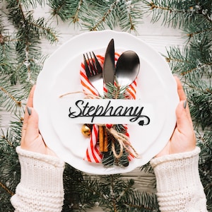 May include: A white plate setting with silverware, a red and white striped napkin, and a name card that reads "Stephany". The setting includes a fork, knife, and spoon, with a sprig of evergreen and a small gold bell. The plate is held by a person wearing a white sweater.