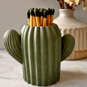 May include: A green cactus-shaped match holder filled with matches. The match holder has a textured surface and two side arms. The matches have dark green tips and are standing upright. A vase with dried flowers is in the background.