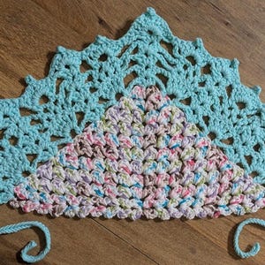 May include: A crocheted doily with a light blue outer edge and a multi-colored center. The doily has a scalloped edge and is made with a delicate stitch.