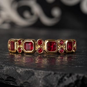 May include: A gold ring with a repeating pattern of red gemstones. The ring features a design of square and round-cut stones, set in a polished gold band. The gemstones are a deep red, creating a contrast against the gold. The ring is displayed on a dark surface.