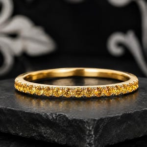 Könnte beinhalten: Ein dünner Goldring mit einer durchgehenden Reihe kleiner, runder, gelber Edelsteine in einer gemeinsamen Krappenfassung. Das Metall hat eine polierte Oberfläche und die Steine sind in Größe und Farbe einheitlich, was für Glanz sorgt.