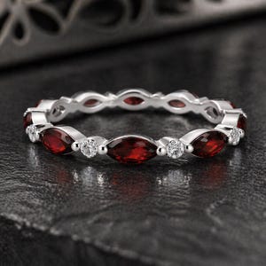 May include: A silver ring with alternating marquise-cut red gemstones and small round white gemstones. The ring is set against a dark, textured surface, highlighting the sparkle of the stones.