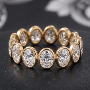 May include: A gold eternity band featuring a continuous row of oval-cut clear stones set in individual bezel settings. The ring is circular, showcasing a repeating pattern of polished gold metal frames surrounding each faceted white gemstone.