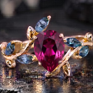 Peut inclure: Une bague en or avec une pierre précieuse centrale en forme de poire d'un rose profond. L'anneau présente un motif de vigne, agrémenté de plusieurs petites pierres bleues taille marquise disposées pour évoquer des feuilles sur une branche.