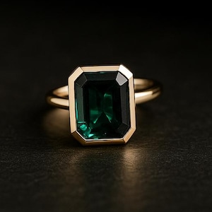 Peut inclure: Une bague en or ornée d'une grande pierre précieuse verte émeraude. La pierre est sertie dans un sertissage en lunette en or, et le corps de la bague est en or poli simple. La bague est présentée sur une surface sombre.