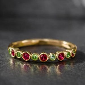 May include: A gold band ring with alternating red and green gemstones. The ring is set with small, round gemstones, creating a colorful and elegant design. The ring is displayed on a dark surface.