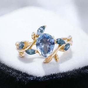 May include: A gold ring with a pear-shaped blue gemstone surrounded by smaller blue gemstones and gold leaves.