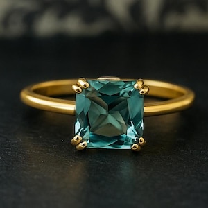 Puede incluir: Un anillo de oro con una piedra preciosa cuadrada de color verde azulado. La piedra preciosa está engastada en un engaste de cuatro puntas. El anillo está sobre una superficie oscura, con un fondo borroso con patrones.