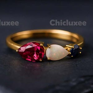 May include: A gold ring featuring three pear-shaped gemstones: a deep red ruby, a white opal, and a dark blue sapphire. The ring is set against a dark background, highlighting the vibrant colors of the stones.