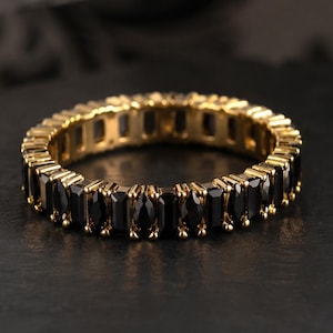 May include: A gold-toned ring with a continuous band of black rectangular stones. The ring is set on a dark, textured surface, with a blurred background featuring a black and white pattern. The ring's design is symmetrical and elegant.