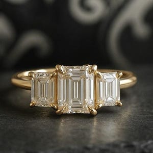 Könnte beinhalten: Ein goldener Ring mit drei Diamanten im Smaragdschliff. Die Diamanten sind in einer Reihe angeordnet, wobei der Mittelstein etwas größer ist als die Seitensteine. Der Ring liegt auf einer dunklen Oberfläche.
