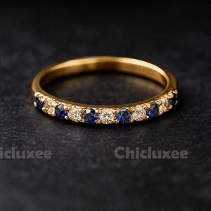 May include: A gold band ring set with alternating blue and white gemstones. The ring is displayed on a dark surface, showcasing its elegant design. The ring is a piece of jewellery.