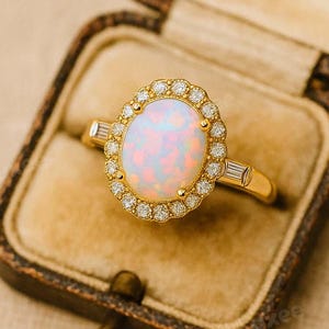 May include: A gold ring with an oval opal center stone, surrounded by a halo of small diamonds. The ring is set in a gold band and is displayed in a vintage-style jewelry box. The opal has a soft, iridescent glow.