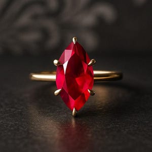 May include: A gold ring featuring a large, marquise-cut, deep red gemstone. The gemstone is held in place by four gold prongs. The ring band is a thin, polished gold. The background is a dark, textured surface.