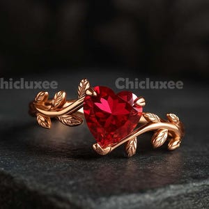 May include: A rose gold ring featuring a heart-shaped, vibrant red gemstone. The ring's band is adorned with delicate leaf-like details, creating an elegant and romantic design. The ring is set against a dark background.