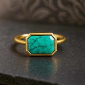 May include: A gold ring featuring a rectangular turquoise stone. The stone is set in a gold bezel and has a matrix of dark veins. The ring band is also gold. The ring is displayed on a dark gray surface.